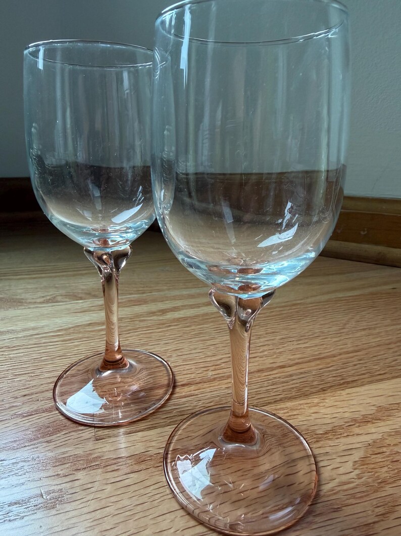 May include: Two clear glass wine glasses featuring tall, transparent bowls and stems with a soft pink tint. Each glass has a wide, circular base that matches the pink color of the stem, providing a stable foundation for the upright drinking vessel.