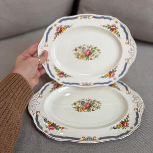 May include: Two oval, off-white ceramic plates with floral designs. Each plate features a central bouquet of colorful flowers and a border of blue ribbon and additional floral accents. The plates are held against a gray background.