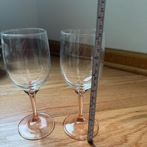 May include: Two clear glass wine glasses with pink stems and bases. A measuring tape placed next to the right glass indicates a total height of approximately 9.5 inches. The glasses feature a smooth, rounded bowl and a slender, colored stem.