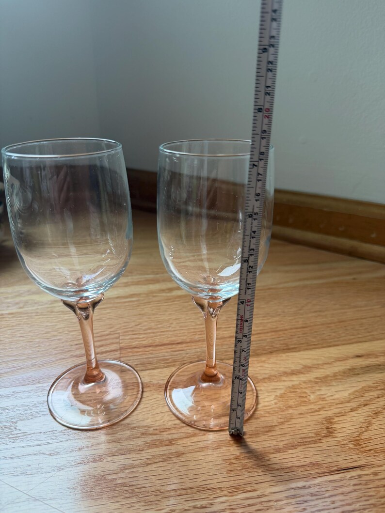 May include: Two clear glass wine glasses with pink stems and bases. A measuring tape placed next to the right glass indicates a total height of approximately 9.5 inches. The glasses feature a smooth, rounded bowl and a slender, colored stem.