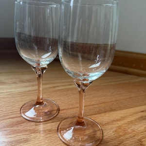May include: Two clear glass wine glasses featuring stems and circular bases tinted in a soft pink hue. The bowls of the glasses are transparent and smooth, while the stems transition from the clear bowl into a delicate pink glass construction.