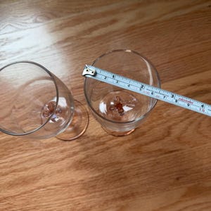 May include: Two clear glass flutes with pink-tinted stems and bases. A white measuring tape is placed across the rim of one flute, showing a diameter of approximately 2.5 inches. The glasses have a smooth, transparent bowl and a decorative base.