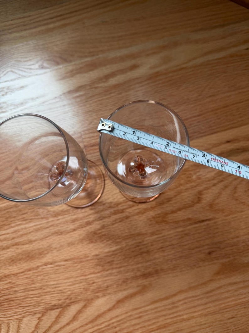 May include: Two clear glass flutes with pink-tinted stems and bases. A white measuring tape is placed across the rim of one flute, showing a diameter of approximately 2.5 inches. The glasses have a smooth, transparent bowl and a decorative base.