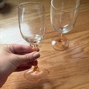 May include: Two clear glass wine glasses featuring a distinct light pink or peach-colored stem and base. The stems have a decorative, textured knot detail just below the bowl of the glass. The glasses have a smooth, rounded rim and a clear bowl.