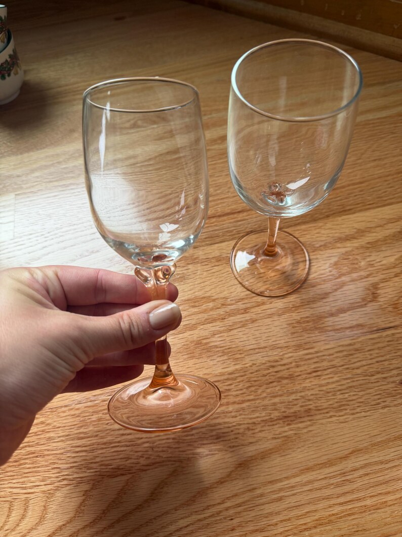 May include: Two clear glass wine glasses featuring a distinct light pink or peach-colored stem and base. The stems have a decorative, textured knot detail just below the bowl of the glass. The glasses have a smooth, rounded rim and a clear bowl.