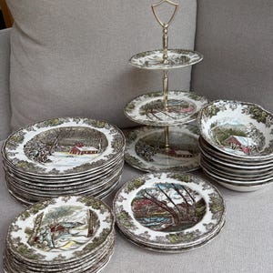 May include: A collection of vintage ceramic dinnerware featuring a winter landscape design. The set includes plates, bowls, and a tiered serving stand. The plates and bowls have a brown and green border and a winter scene in the center.