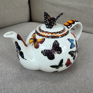 May include: A white ceramic teapot decorated with various colorful butterflies in shades of orange, black, blue, and green. The lid features a three-dimensional butterfly handle, and the handle of the teapot also displays a butterfly wing pattern.