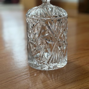 May include: A clear, cut-glass jar with a lid. The jar has a cylindrical shape with a diamond and starburst pattern. The lid has a faceted knob. The jar is sitting on a wooden surface.