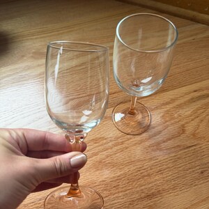 May include: Two clear glass wine glasses featuring delicate pink stems and circular bases. The bowls are transparent and smooth, while the stems transition into a soft pink hue that extends to the base, providing a subtle color contrast.