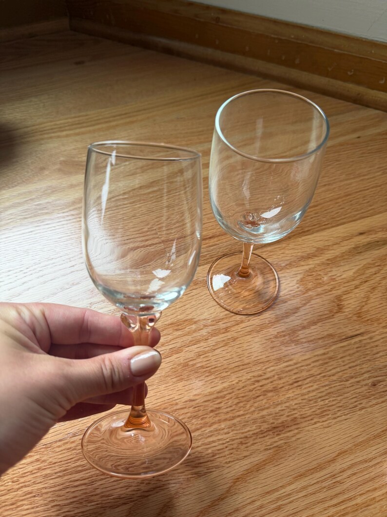 May include: Two clear glass wine glasses featuring delicate pink stems and circular bases. The bowls are transparent and smooth, while the stems transition into a soft pink hue that extends to the base, providing a subtle color contrast.