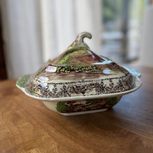 May include: A ceramic lidded serving dish featuring a brown and green transferware landscape pattern. The rectangular base has rounded corners and side handles, while the lid includes a curved handle and a detailed pastoral scene.