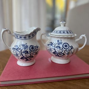 May include: A white ceramic creamer and a lidded sugar bowl featuring a blue floral pattern. Both pieces have a scalloped rim, a pedestal base, and ornate handles. The blue design depicts stylized flowers and leaves on the body and lid.