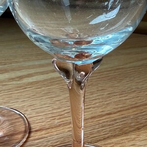 May include: A clear glass wine goblet featuring a delicate pink-tinted stem and base. The bowl is transparent and rounded, transitioning into a sculpted, flared neck that connects to the slender, colored stem and a circular, flat foot.