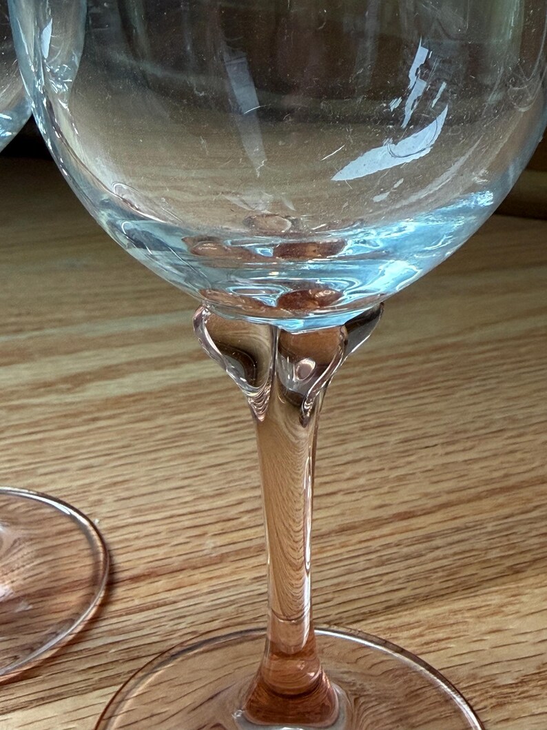 May include: A clear glass wine goblet featuring a delicate pink-tinted stem and base. The bowl is transparent and rounded, transitioning into a sculpted, flared neck that connects to the slender, colored stem and a circular, flat foot.