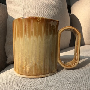 May include: A ceramic mug featuring a cylindrical body with a vertical, drip-style glaze in shades of honey, amber, and cream. The mug has a wide, rectangular-shaped handle that extends from the side, maintaining the same warm, glossy finish.