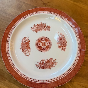 May include: A white ceramic plate with a red floral design. The plate has a red border with a geometric pattern. The center of the plate features a red floral design. The plate is on a wooden surface.