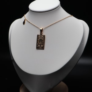 May include: A gold-coloured necklace with a rectangular pendant featuring an engraved design. The necklace is displayed on a white bust against a black background. The pendant has intricate details.