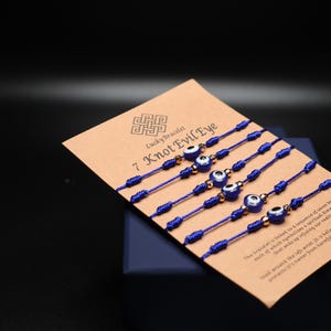 Könnte beinhalten: Ein Set aus sieben blauen Armbändern mit einem bösen Blick und goldfarbenen Akzenten, präsentiert auf einer braunen Karte. Die Karte trägt den Text "Lucky Bracelet 7 Knot Evil Eye". Die Armbänder sind geknotet.