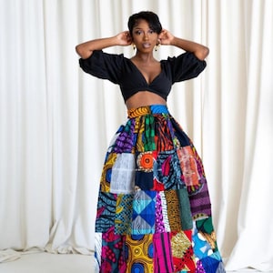 May include: A black crop top paired with a vibrant, patchwork maxi skirt. The skirt features a variety of colorful African-inspired fabric patterns, including geometric shapes and floral designs. The outfit is set against a white curtain backdrop.