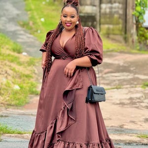 May include: A woman wearing a long, flowing, brown dress with a wrap-style bodice and ruffled skirt. The dress has short, puffed sleeves. She carries a small, black purse with a chain strap. Her hair is styled in braids and a bun.