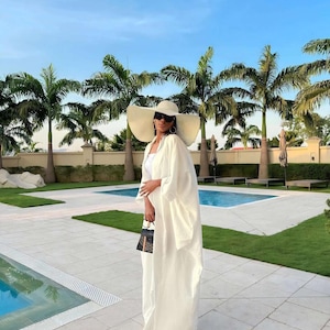 May include: A woman wearing a white outfit, including a wide-brimmed hat and a long, flowing robe, stands near a swimming pool. She is also wearing sunglasses and carrying a small black handbag. Palm trees and a blue sky provide a backdrop.