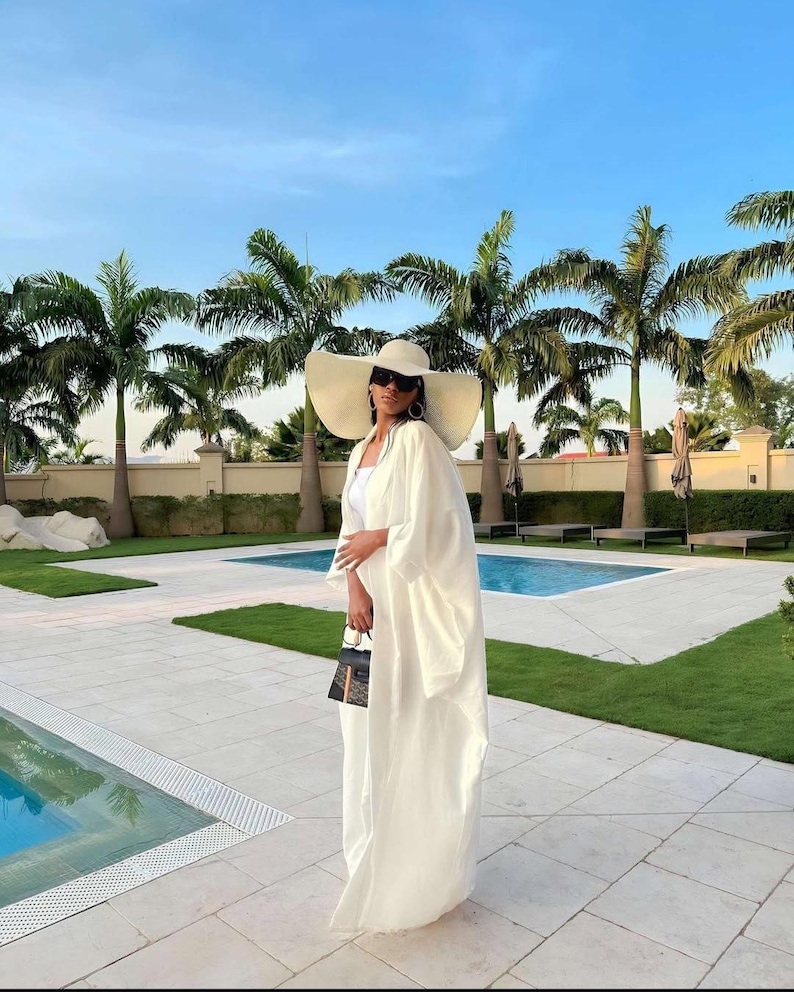 May include: A woman wearing a white outfit, including a wide-brimmed hat and a long, flowing robe, stands near a swimming pool. She is also wearing sunglasses and carrying a small black handbag. Palm trees and a blue sky provide a backdrop.