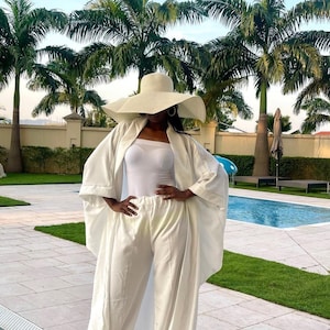 May include: A person wearing a white outfit, including a wide-brimmed hat, a long jacket, a tank top, and wide-leg pants. The outfit is all white. The background includes palm trees, a swimming pool, and a manicured lawn.