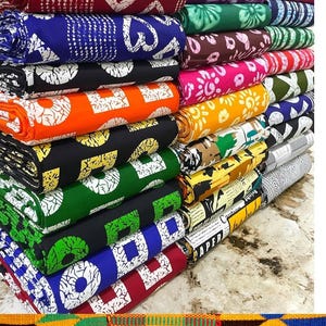 Pode incluir: Uma pilha de rolos de tecido com estampas tribais coloridas. Os tecidos apresentam vários padrões em cores como preto, vermelho, azul, laranja e verde. O texto na imagem diz "Wholesale (25 pieces) Tribal prints 100% cotton".
