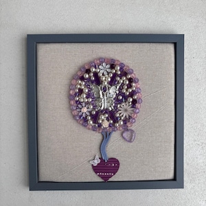 Può includere: Un'opera d'arte da parete incorniciata con una disposizione circolare di perline viola e trasparenti, accenti floreali bianchi e una farfalla argentata centrale. Un cordino intrecciato blu pende verso un cuore viola con una piccola farfalla bianca.