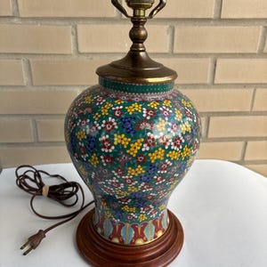 Op de afbeelding: Een tafellamp met een bolvormige, groene basis voorzien van een dicht, veelkleurig bloemenpatroon. De lamp heeft een messingkleurige metalen hals en fitting, een ronde voet van donker hout en een bruin elektrisch snoer met een stekker.