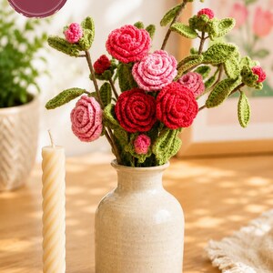Pode incluir: Um arranjo de flores em crochê com rosas vermelhas e cor-de-rosa, folhas e caules verdes num vaso de cerâmica bege, acompanhado por uma vela cónica torcida de cor creme num suporte de metal dourado. A imagem inclui uma etiqueta PDF PATTERN.