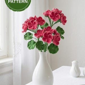 May include: A white ceramic vase holds a bouquet of crocheted flowers. The flowers are a vibrant pink, with green crocheted leaves. The vase sits on a white tablecloth. A green circle with the text "PDF PATTERN" is in the upper left.