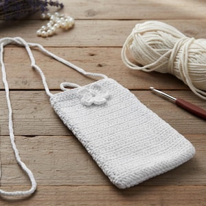 Könnte beinhalten: Eine weiße, gehäkelte Handytasche mit Trageriemen und Blumenverzierung. Die Tasche liegt auf einer Holzoberfläche, im Hintergrund befinden sich ein Wollknäuel, eine Häkelnadel und Lavendelzweige.
