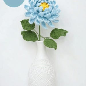Könnte beinhalten: Eine gehäkelte blaue Chrysantheme mit gelber Mitte und grünen Blättern in einer weißen Vase. Die Vase hat ein dekoratives Muster. Das Bild enthält einen blauen Kreis mit dem Text "PDF PATTERN".
