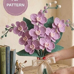May include: A handcrafted bouquet of crocheted lavender orchids with green leaves and stems, tied with a cream-colored ribbon. The bouquet features a PDF pattern label.