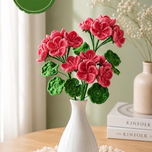 May include: A crochet PDF pattern featuring a bouquet of vibrant pink geranium flowers with textured green leaves. The flowers are arranged in a smooth white ceramic vase, showcasing intricate yarn stitching and detailed petal structures.