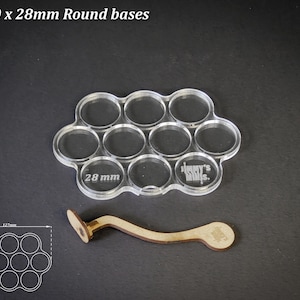 Op de afbeelding: Transparante acryl basis houder voor 10 ronde bases, elk met een diameter van 28 mm, met de tekst "28 mm" en "Jimmy's Mills". Een houten handvat is ook inbegrepen. De tekst "For 10 x 28mm Round bases" staat bovenaan.
