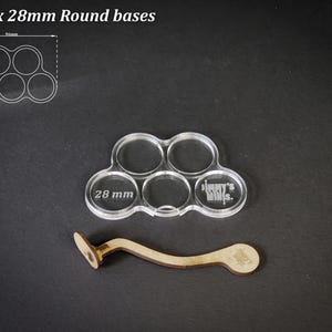 Puede incluir: Base de acr&iacute;lico transparente para 5 bases redondas de 28 mm, con un mango de madera. La base tiene cinco recortes circulares, cada uno de 28 mm de di&aacute;metro. El texto en la base dice "Jimmy's Minis". El mango de madera es curvo.