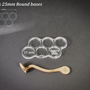 May include: Clear acrylic base holder for six 25mm round bases, with a wooden handle. The holder has six circular openings. The text "25 mm" is visible on the acrylic. The wooden handle is light brown.