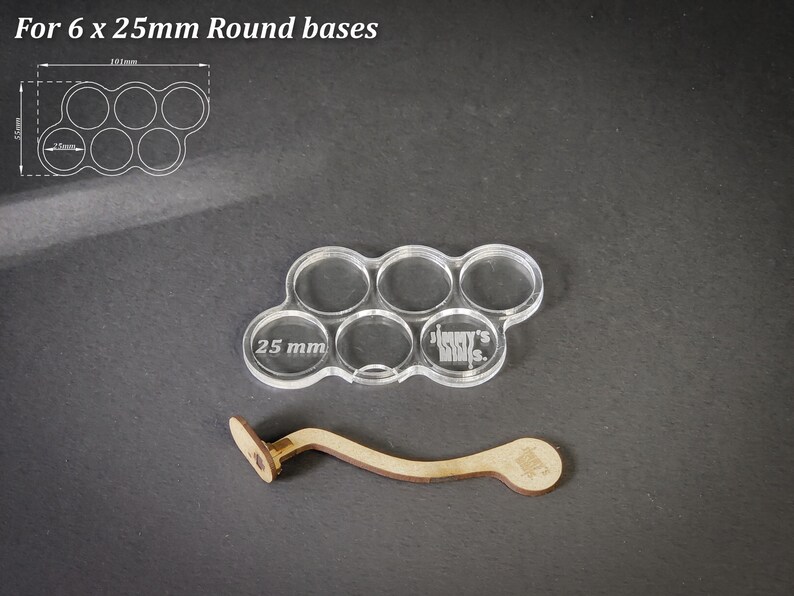 May include: Clear acrylic base holder for six 25mm round bases, with a wooden handle. The holder has six circular openings. The text "25 mm" is visible on the acrylic. The wooden handle is light brown.