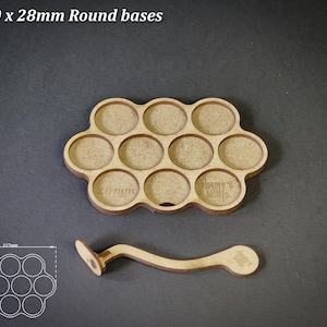 May include: A light brown wooden tray with ten circular recesses, each for 28mm round bases, and a matching handle. The tray has the text "For 10 x 28mm Round bases" at the top. The tray is 127mm x 65mm.