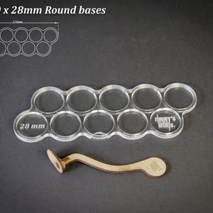 May include: Clear acrylic base holder for 10 miniature figures with 28 mm round bases. Includes a wooden tool for easy placement. The text "For 10 x 28mm Round bases" is at the top. The holder is 17.4cm long and 6.1cm wide.