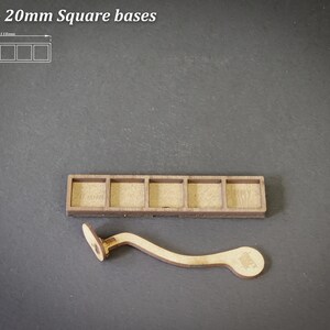 Puede incluir: Soporte de base cuadrada de madera con cinco compartimentos y una herramienta de madera curvada. El texto "For 5 - 20mm Square bases" est&aacute; en la parte superior. El soporte mide aproximadamente 11,1 cm de largo.