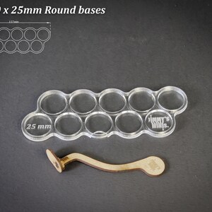 May include: Clear acrylic base for 10 miniature figures with 25 mm round bases. Includes a wooden handle. The text "For 10 x 25mm Round bases" is at the top. The base has 10 circular cutouts.