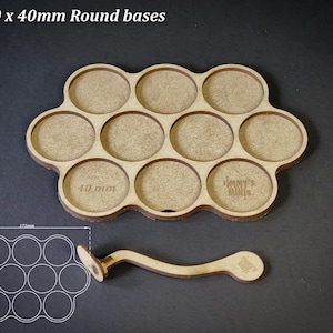 May include: A wooden base designed to hold ten 40mm round bases, with a matching handle. The base is light brown with circular cutouts and the text "For 10 x 40mm Round bases". The handle has a circular end and a curved design.