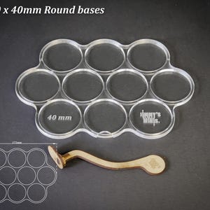 Op de afbeelding: Transparante acryl basis houder voor 10 ronde bases, elk met een diameter van 40 mm. Inclusief een houten gereedschap. De tekst "For 10 x 40mm Round bases" staat bovenaan. Een diagram toont de afmetingen.