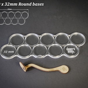 May include: Clear acrylic base holder for 10 x 32mm round bases, with a wooden tool. The holder has 10 circular cutouts, each 3.2 cm in diameter. The text "For 10 x 32mm Round bases" is at the top.