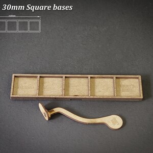 Puede incluir: Base de madera para 5 miniaturas cuadradas, con un asa curva. La base tiene cinco ranuras rectangulares, cada una de 30 mm cuadrados. El texto "For 5 - 30mm Square bases" est&aacute; en la parte superior. El asa facilita el transporte.