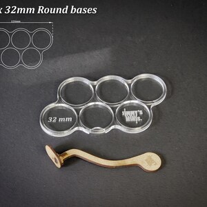 May include: Clear acrylic base holder for six 32mm round bases, with a wooden handle. The holder has six circular openings, each labeled with "32 mm". The text "For 6 x 32mm Round bases" is in the upper left corner.