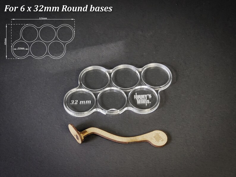 May include: Clear acrylic base holder for six 32mm round bases, with a wooden handle. The holder has six circular openings, each labeled with "32 mm". The text "For 6 x 32mm Round bases" is in the upper left corner.